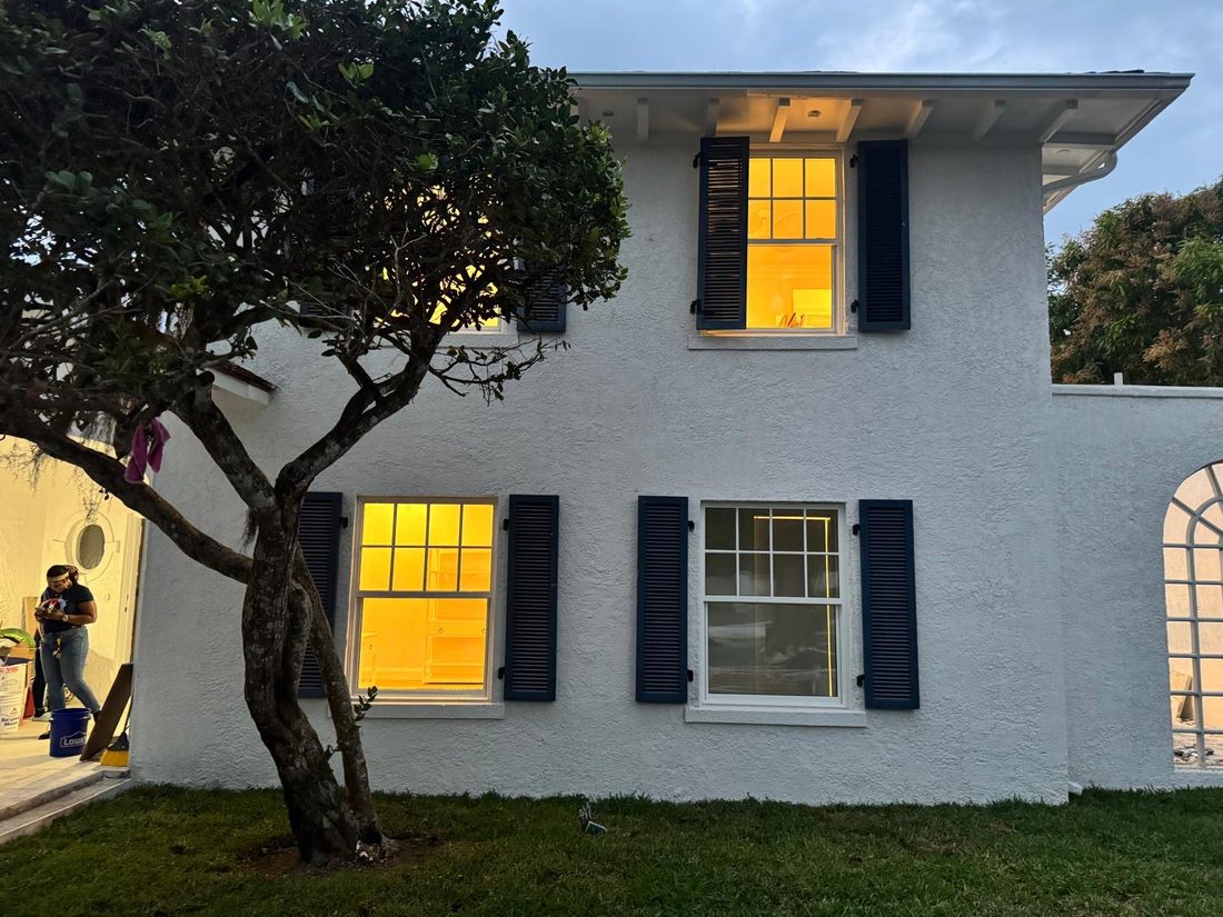 White stucco elevation at Lyford Cay with navy shutters, lit from inside at dusk.